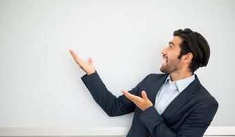 Portrait of smiling young caucasian businessman wearing dark suit and presenting copy space on his palms near with concrete wall. The man feeling happy and excited. photo