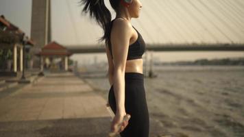 Dolly Shot, una mujer asiática blanca con un sostén deportivo negro y pantalones negros, está haciendo un entrenamiento de salto de cuerda. en medio del río por la mañana o por la tarde video