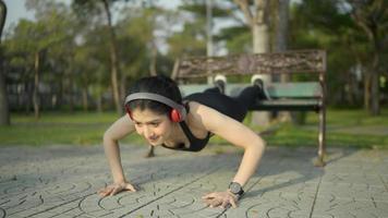vídeo em câmera lenta. uma mulher asiática branca vestindo um sutiã esportivo preto. calças pretas e fones de ouvido vermelhos fazendo exercício com flexões no parque. video