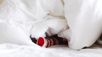 children feet in red white striped christmas wool knitted socks under white fluffy blanket. cool cozy winter. heating, energy crisis and consumption concept video