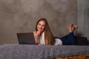 mujer sonriente acostada boca abajo en la cama con una laptop frente a ella y mirando a la cámara foto