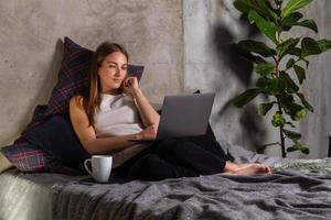 mujer sentada en la cama con una laptop en el regazo y una taza blanca a un lado foto