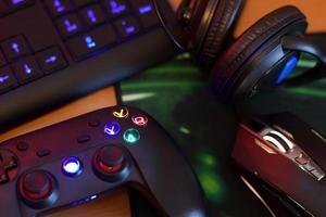 Modern gamepad and gaming mouse lies with keyboard and headphones on table in dark playroom scene. Cheating in online video games concept photo