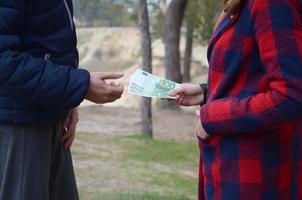 Girl transfers euro bills to the hands of a young guy in forest. Concept of robbery or illegal deal transaction photo