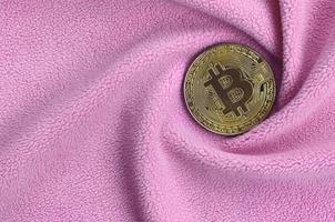 The golden bitcoin lies on a blanket made of soft and fluffy light pink fleece fabric with a large number of relief folds. The shape of the folds resembles a fan from a video card cooler photo