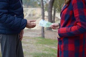 Girl transfers euro bills to the hands of a young guy in forest. Concept of robbery or illegal deal transaction photo