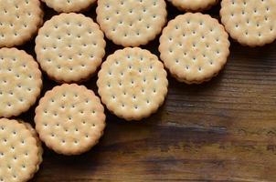 A round sandwich cookie with coconut filling lies in large quantities on a brown wooden surface. Photo of edible treats on a wooden background with copy space