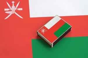 Oman flag is pictured on a matchbox that lies on a large flag photo