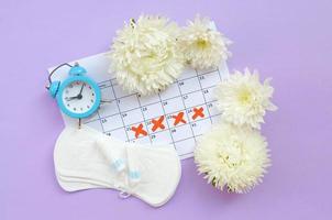 Menstrual pads and tampons on menstruation period calendar with blue alarm clock and white flowers photo