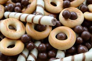 A lot of crispy sweet tubules, chocolate melting balls and yellow bagels lie on a wooden surface. Close up view photo