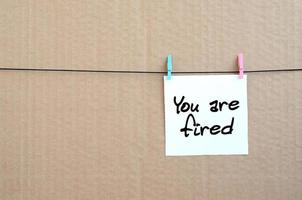 You are fired. Note is written on a white sticker that hangs with a clothespin on a rope on a background of brown cardboard photo