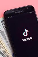 Tiktok application on samsung smartphone screen and dollar bills. TikTok is a popular video-sharing social networking service owned by ByteDance photo