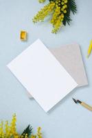 White sheet of paper for text copy space surrounded by spring mimosa, notepad, yellow pencil, watercolors in cuvettes on a blue background. Workplace of a blogger, artist, or calligrapher photo