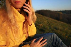 Blonde woman in yellow sweater and blue jeans talking on the phone and sitting on hill in orange sunset light. Cropped image, without face, unrecognizable, only lips are visible. Autumn warm mood. photo