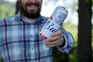 sumy, ucrania - 01 de agosto de 2021 un joven levanta la lata de cerveza budweiser bud y muestra el logotipo de bud en el fondo borroso del río y los árboles. budweiser es una de las marcas de cerveza más populares en los estados unidos foto