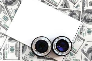 Two photographic lenses and white notebook lie on the background of a lot of dollar bills. Space for text photo