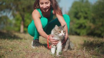 A beautiful athletic woman in a sporty turquoise overalls walks with her fluffy cat on a leash in the forest on a green lawn. Cute kitten walks in the park with the owner. Pet care. Slow motion. video