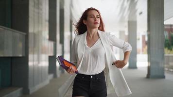 mooi zelfverzekerd bedrijf vrouw met rood haar- in mode kleren wandelen naar werk in de buurt kantoor. jong dame genieten van geslaagd zakelijke carrière. elegant vrouw ondernemer wandelingen binnenstad. langzaam beweging. video