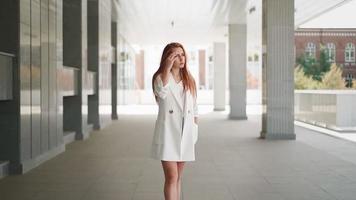 belle femme d'affaires moderne aux longs cheveux roux en manteau blanc à la mode se tient près d'un immeuble de bureaux. femme entrepreneur confiante. jolie dame réussie dans des vêtements à la mode. ralenti. video