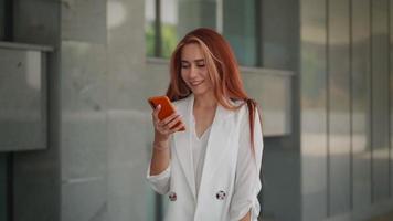 hermosa mujer de negocios sonriendo y usando mensajes de texto de teléfonos inteligentes en la calle. joven dama exitosa junto a la oficina corporativa escribiendo mensajes de texto en el teléfono. mujer empresaria revisando correos electrónicos. camara lenta. video