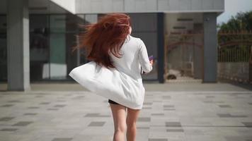 en skön Lycklig kvinna med lång röd hår i en trendig vit täcka promenader och ler, fröjd i henne Framgång och frihet. lekfull lady går dans i stadens centrum. långsam rörelse. video