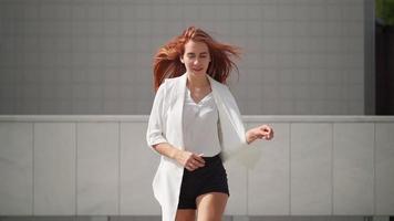 A beautiful woman with long red hair in a white coat runs along the city street. A pretty playful lady is running in casual clothes. Slow motion. video