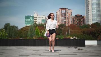 una hermosa mujer con el pelo largo y rojo con un abrigo blanco corre por la calle de la ciudad. una dama bastante juguetona corre con ropa informal. camara lenta. video