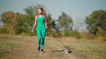 una hermosa mujer atlética con un mono deportivo turquesa camina con su gato esponjoso con una correa en el bosque sobre un césped verde. lindo gatito camina en el parque con el dueño. cuidado de mascotas. camara lenta. video