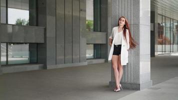 Beautiful confident business woman with red hair in fashion clothes stands near the corporate office. Young lady enjoying successful career. Elegant female entrepreneur downtown. Slow motion. video