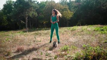 A beautiful athletic woman in a sporty turquoise overalls walks with her fluffy cat on a leash in the forest on a green lawn. Cute kitten walks in the park with the owner. Pet care. Slow motion. video