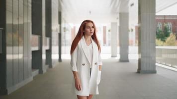 mooi modern bedrijf vrouw met lang rood haar- in modieus wit jas staat in de buurt een kantoor gebouw. zelfverzekerd vrouw ondernemer. aantrekkelijk geslaagd dame in modieus kleren. langzaam beweging. video