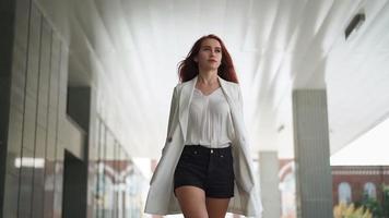 Beautiful confident business woman with red hair in fashion clothes walk to work near office. Young lady enjoying successful corporate career. Elegant female entrepreneur walks downtown. Slow motion. video