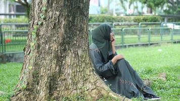 mujer joven en hiyab oscuro sentado bajo un árbol en la hierba video