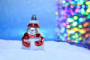 Snowman with a red scarf in the snow and blue sky from behind and multicolored bokeh. Blurred background. Merry Christmas and new year card. Copy space photo