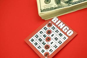 tablero de bingo rojo o naipe para ganar fichas y pila de billetes de dólar. Tarjeta de bingo clásica americana o canadiense de cinco a cinco sobre fondo rojo. foto