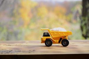 A small yellow toy truck is loaded with a stone of white salt. A car on a wooden surface against a background of autumn forest. Extraction and transportation of salt photo