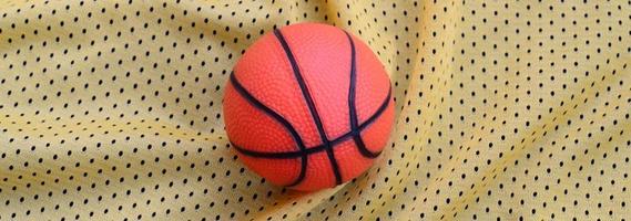 Small orange rubber basketball lies on a yellow sport jersey clothing fabric texture and background with many folds photo