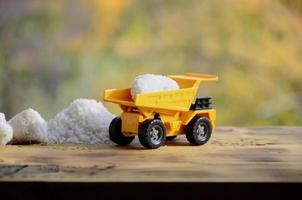 A small yellow toy truck is loaded with a stone of white salt next to a pile of salt. A car on a wooden surface against a background of autumn forest. Extraction and transportation of salt photo