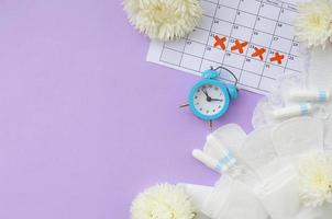 Menstrual pads and tampons on menstruation period calendar with blue alarm clock and white flowers photo