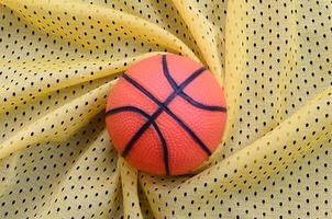 Small orange rubber basketball lies on a yellow sport jersey clothing fabric texture and background with many folds photo