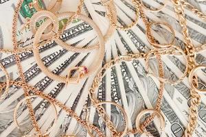 Many expensive golden jewerly rings, earrings and necklaces on big amount of US dollar bills close up. Pawnshop or jewerly shop photo