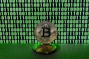 A pair of bitcoins lies on a cardboard surface on the background of a monitor depicting a binary code of bright green zeros and one units on a black background. Low key lighting photo