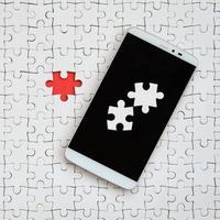 A modern big smartphone with several puzzle elements on the touch screen lies on a white jigsaw puzzle in an assembled state with missing elements photo