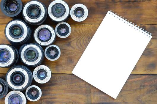 Several photographic lenses and white notebook lie on a brown wooden background. Space for text photo
