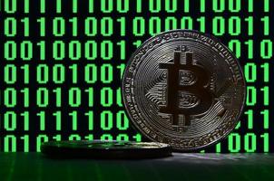 A pair of bitcoins lies on a cardboard surface on the background of a monitor depicting a binary code of bright green zeros and one units on a black background. Low key lighting photo