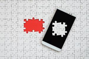 A modern big smartphone with several puzzle elements on the touch screen lies on a white jigsaw puzzle in an assembled state with missing elements photo