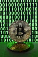 A pair of bitcoins lies on a cardboard surface on the background of a monitor depicting a binary code of bright green zeros and one units on a black background. Low key lighting photo