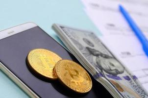 bitcoins se encuentra con los formularios de impuestos, los billetes de cien dólares y el teléfono inteligente en un fondo azul claro. declaración de la renta foto