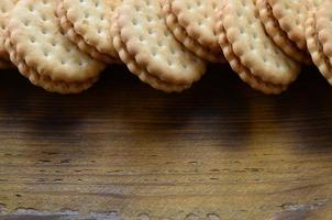 A round sandwich cookie with coconut filling lies in large quantities on a brown wooden surface. Photo of edible treats on a wooden background with copy space