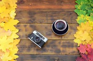 A cup of tea and an old camera among a set of yellowing fallen autumn leaves on a background surface of natural wooden boards of dark brown color photo